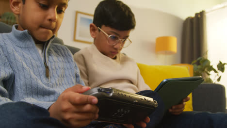 Zwei-Junge-Jungen-Sitzen-Zu-Hause-Auf-Dem-Sofa-Und-Spielen-Spiele-Oder-Streamen-Auf-Ein-Digitales-Tablet-Und-Ein-Handheld-Spielgerät-1